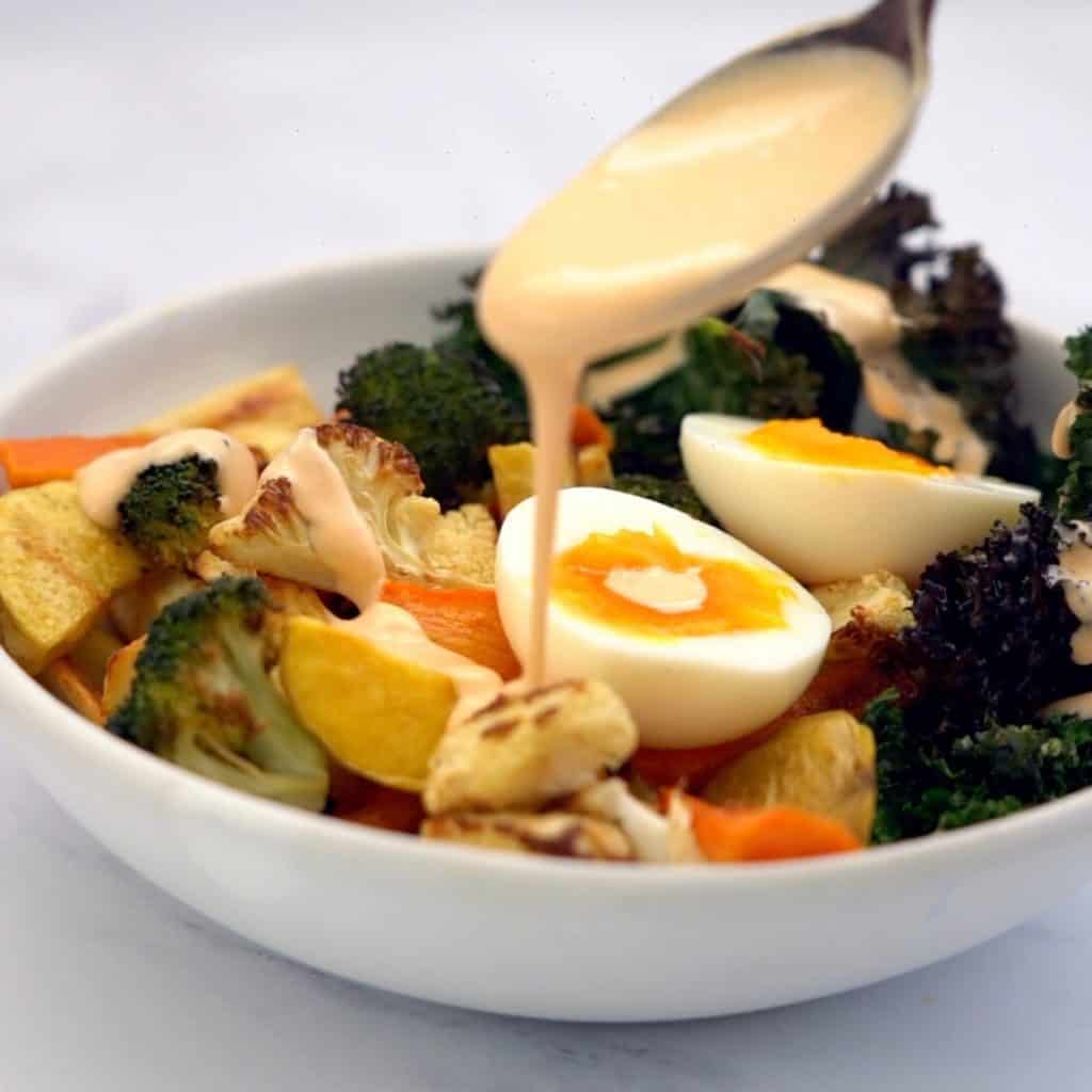 spoonful of aioli being drizzled over a bowl of roast vegetables and hard boiled eggs