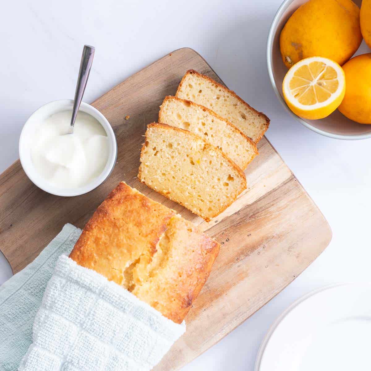 Lemon loaf on a wooding chopping board with a bowl of yoghurt and a bowl of lemons