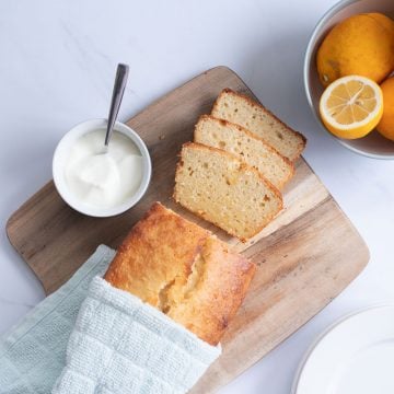 lemon loaf on a wooden chopping board with a bowl of lemons and yoghurt