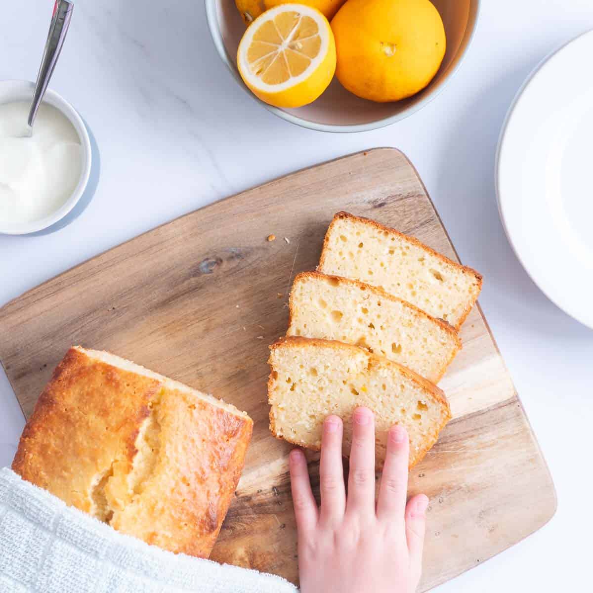 3 Slices of lemon loaf on a wooden chopping board with a child's hand reaching out for a piece