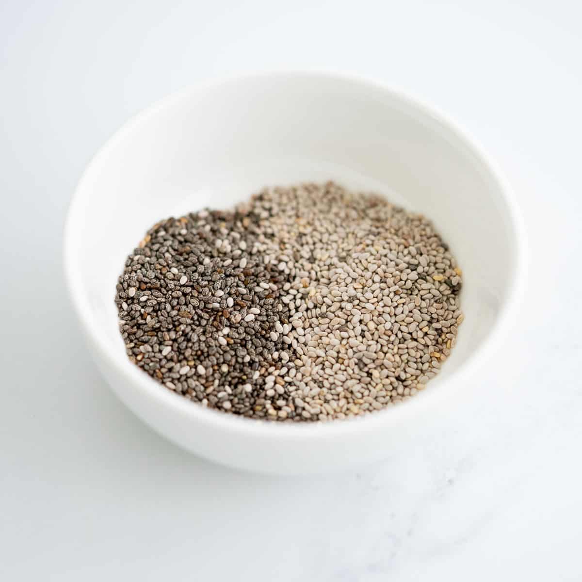 Small white bowl half filled with white chai seeds and half filled with black chia seeds.