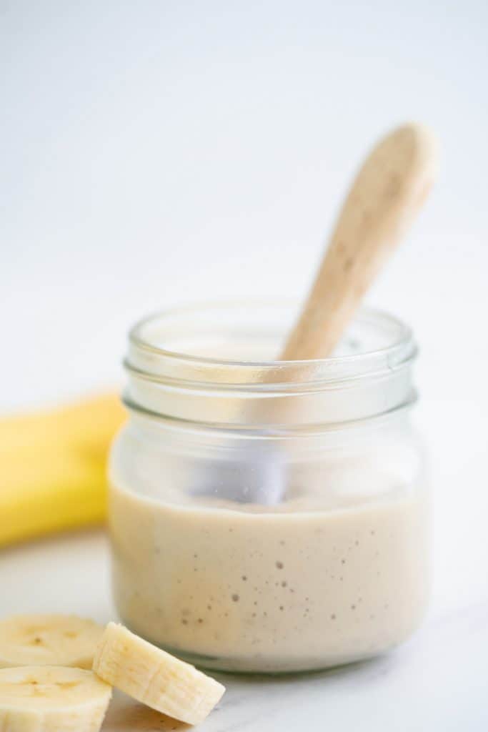 A glass jar filled with pudding with a bamboo handled baby spoon and banana slices. 