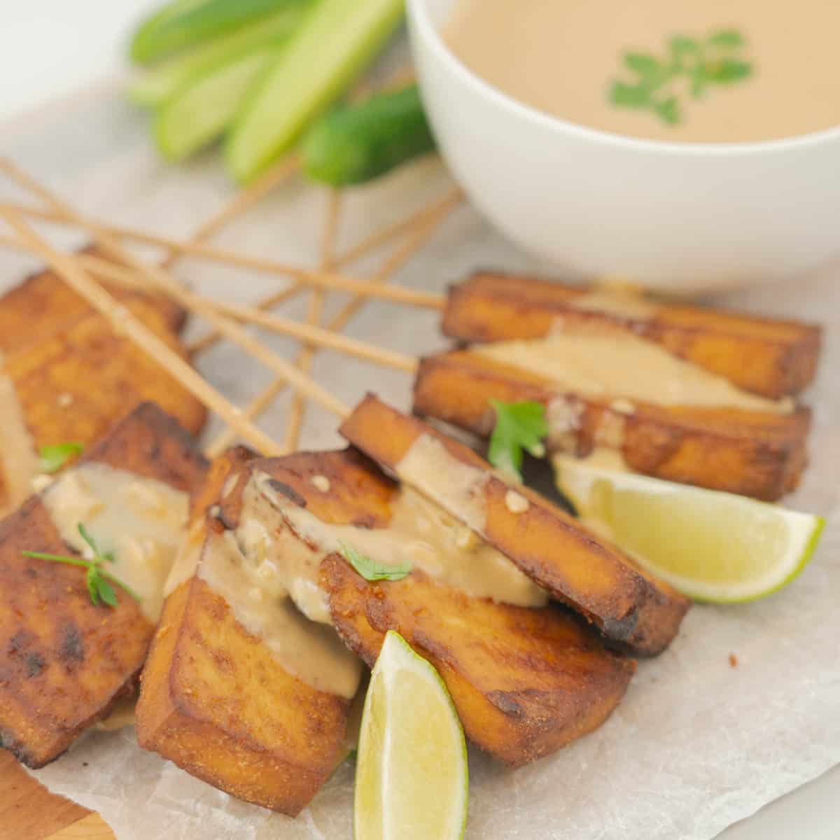 tofu skewers drizzled with satay sauce on a board with lime wedges.