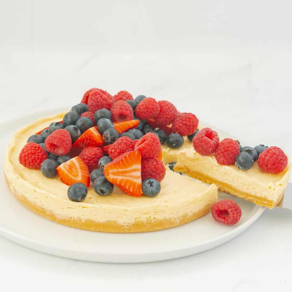 A round cheesecake with biscuit base topped with berries on a large white serving platter, a cake slice is removing one piece.