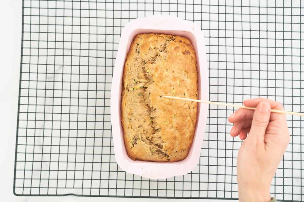 baked banana bread in loaf tin with a woman's hand holding a clean skewer