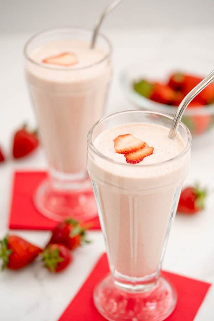 Two strawberry milkshakes in tall glasses topped with strawberry slice with red napkins.