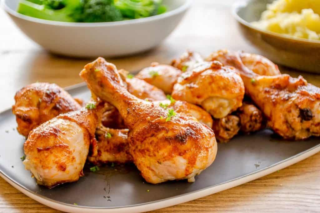 A platter of paprika chicken drumsticks in the foreground with broccoli and mashed potato in the background.