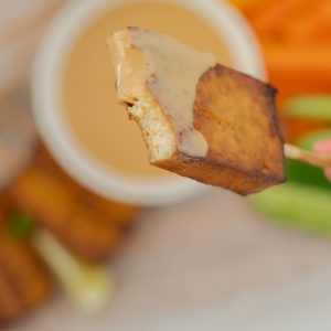 A hand holding a piece of baked tofu on a skewer, a mouthful has been eaten, revealing the creamy white centre of the tofu.