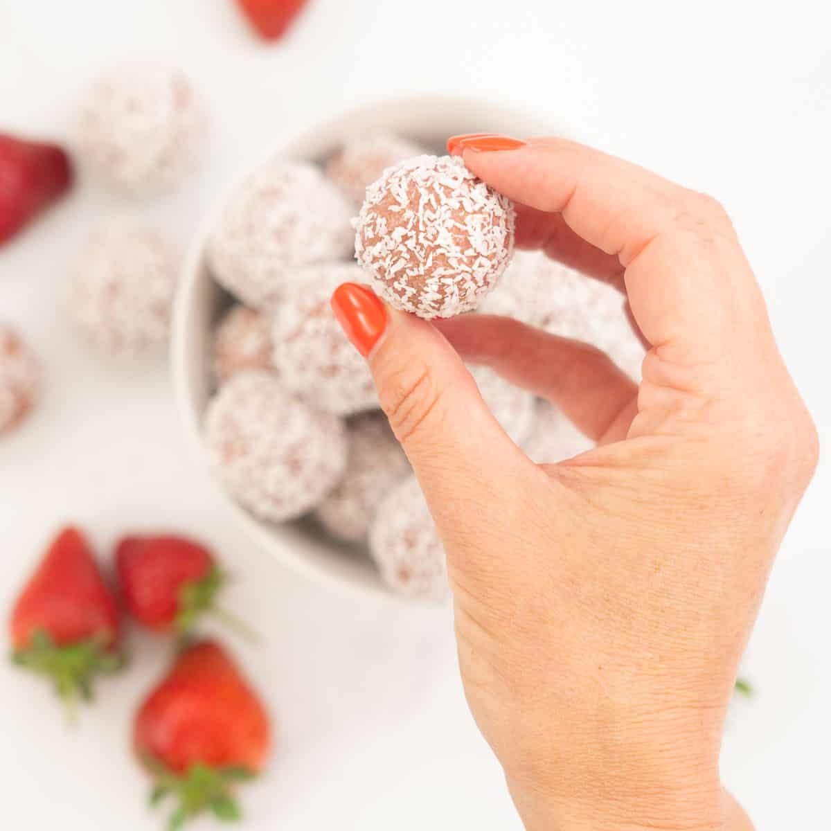 A woman's hand holding a coconut covered strawberry bliss ball.