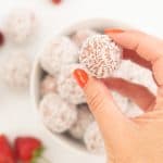 A woman's hand holding a coconut covered strawberry bliss ball.