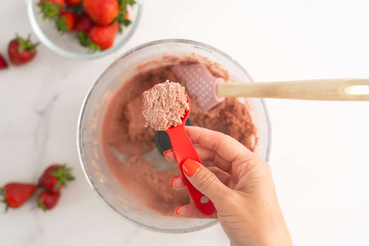 A womans hand scooping a tablespoon of pink strawberry bliss ball mix.