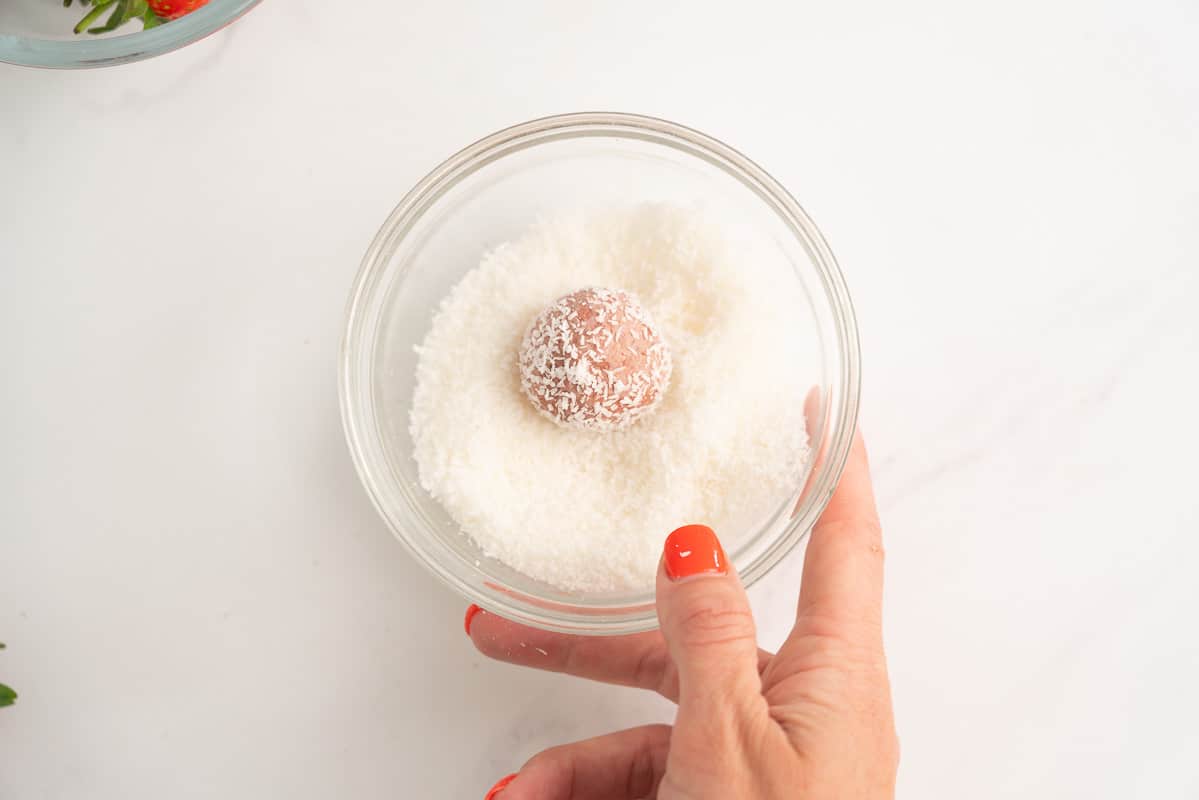 A small bowl filled desiccated coconut, a woman's hand rolling a pink bliss ball in the coconut.