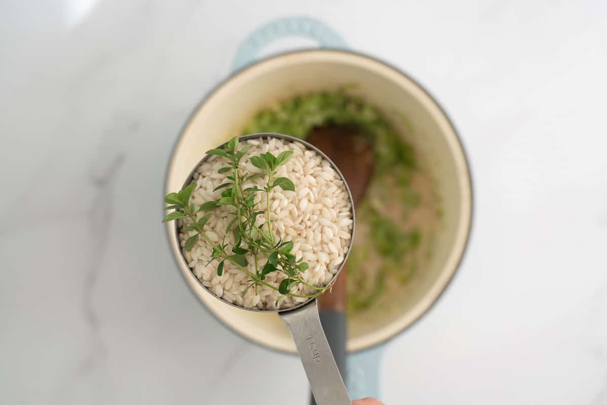 A measuring cup of arborio rice and sprigs of thyme held above a large ceramic saucepan.