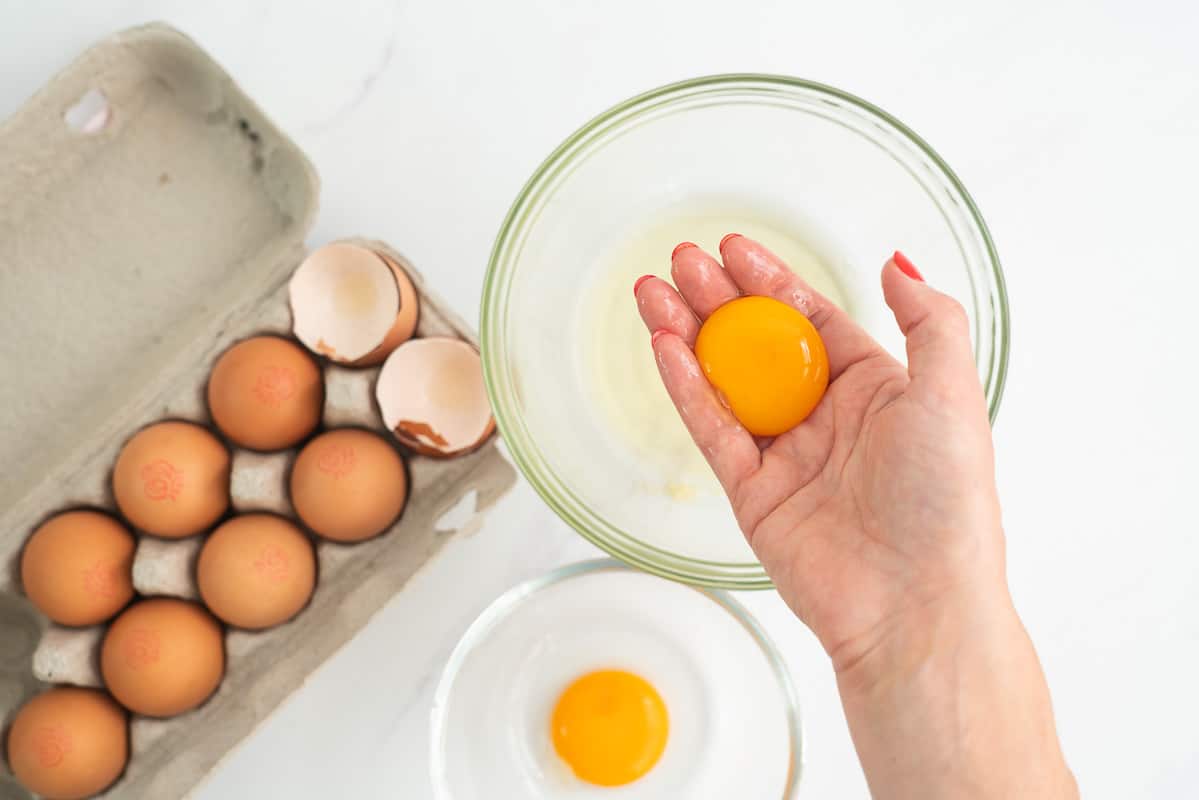 A woman's hand holding an egg yolk above a bowl of egg whites.