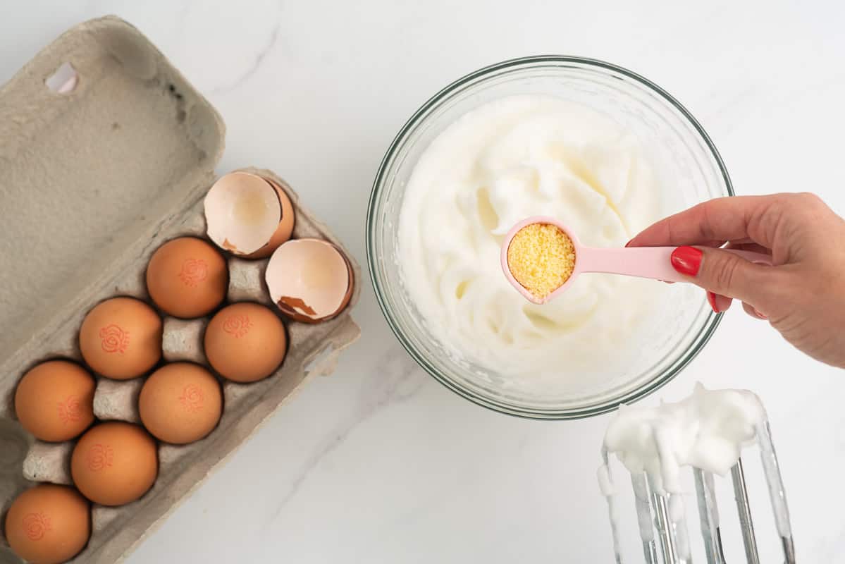 A woman's hand holding a tablespoon of parmesan cheese above a bowl of egg whites.