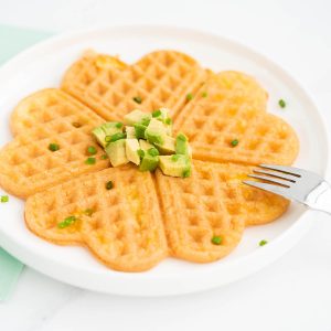 An egg waffle on a white plate garnished with small cubes of avocado and chives.