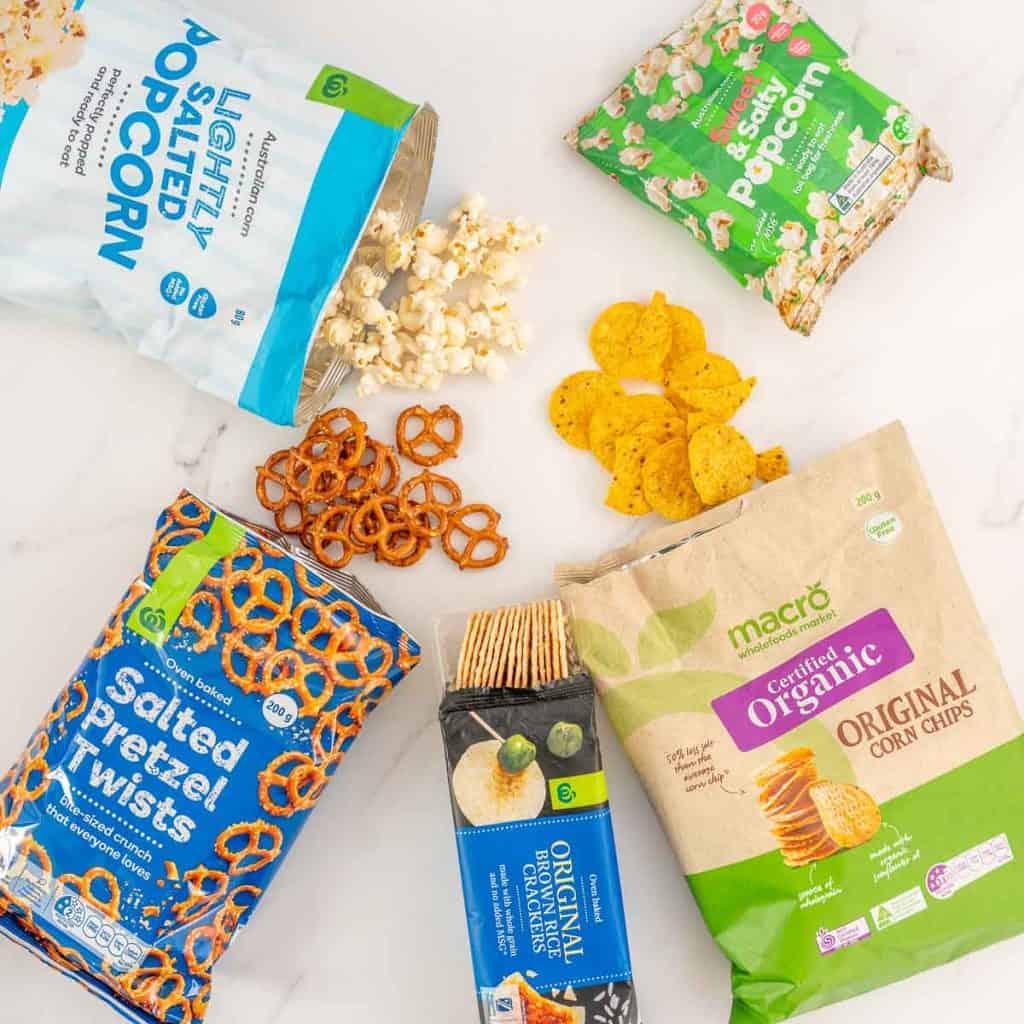 Popcorn, crackers, pretzels, and corn chips laid out on a bench top