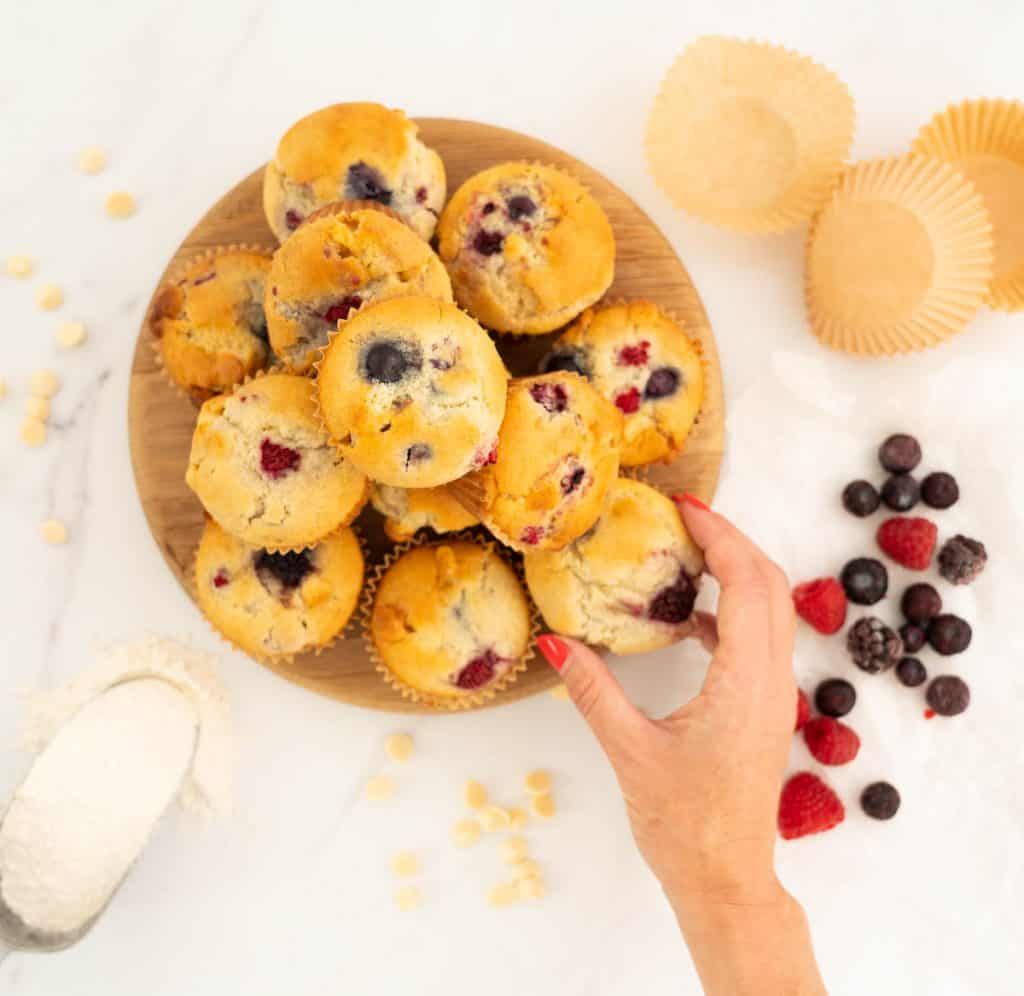 Women's hand reaching for a berry muffin from a wooden tray of a dozen muffins.