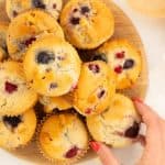 Women's hand reaching for a berry muffin from a wooden tray of a dozen muffins.