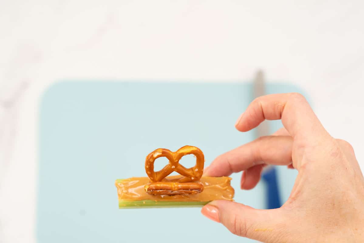 A women's hand holding a celery stick filled with peanut butter and decorated with pretzels.