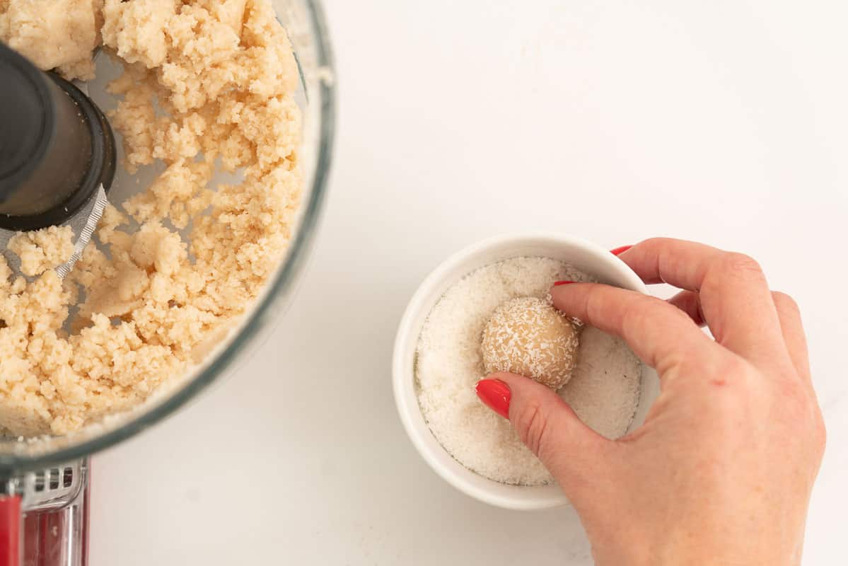 A coconut bliss ball being rolled in a small bowl of desiccated coconut.