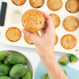 Women's hand holding a feijoa muffin above a bowl of green feijoas.