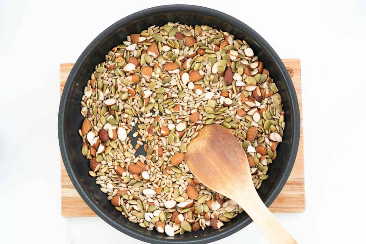 Sunflower seeds, almonds and pumpkin seeds in a large fry pan with a wooden spoon.