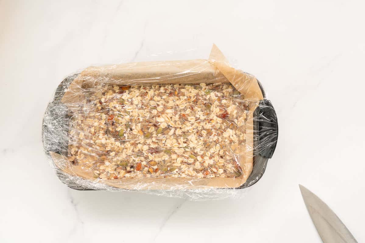 Uncooked oatmeal bread, in a loaf tin wrapped with cling film, ready to be refrigerated.