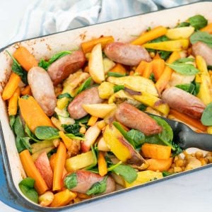 Baked sausages, apples, sweet potato and spinach leaves in a blue ceramic roasting pan.