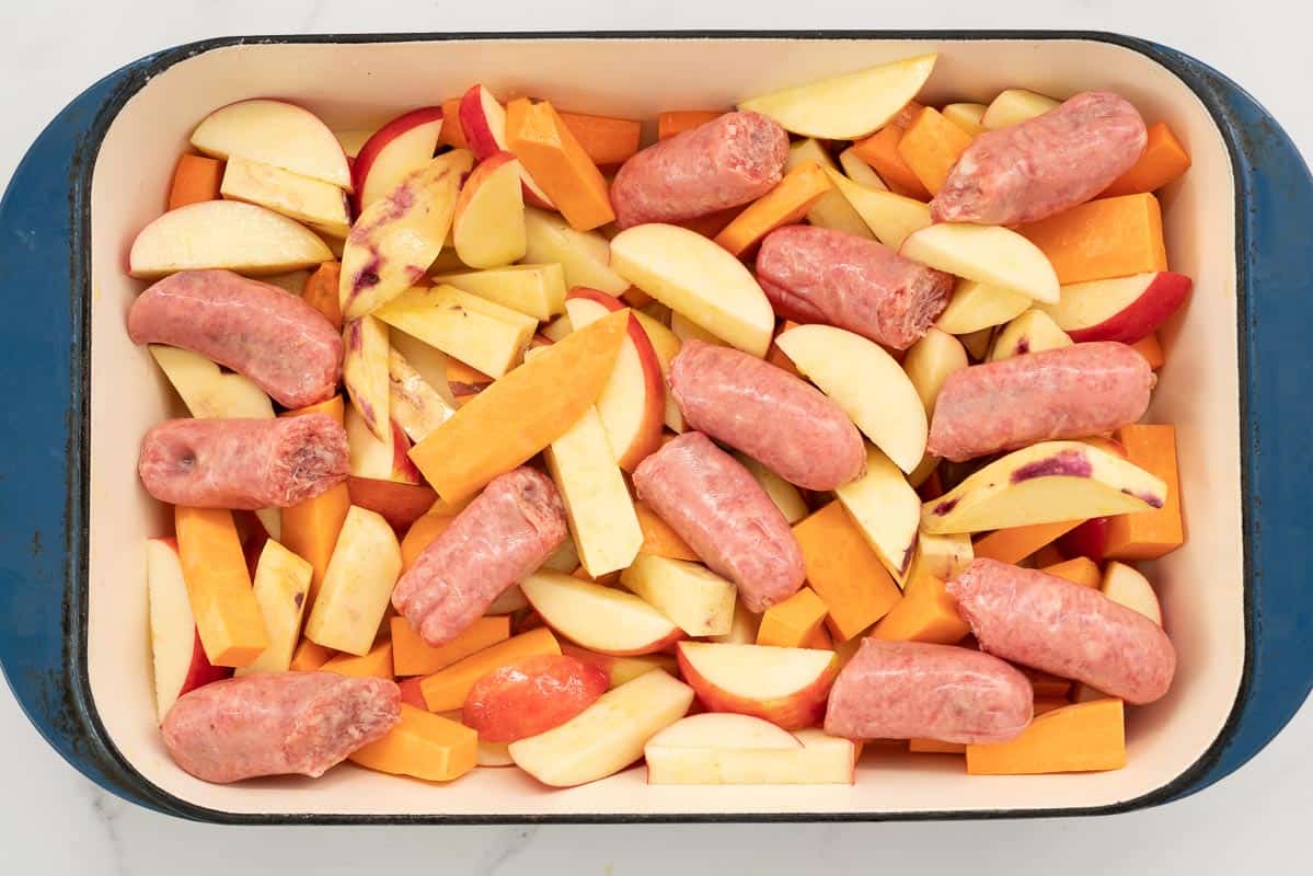 Sliced apples, carrots, sweet potato and sausages in a large blue ceramic roasting pan.