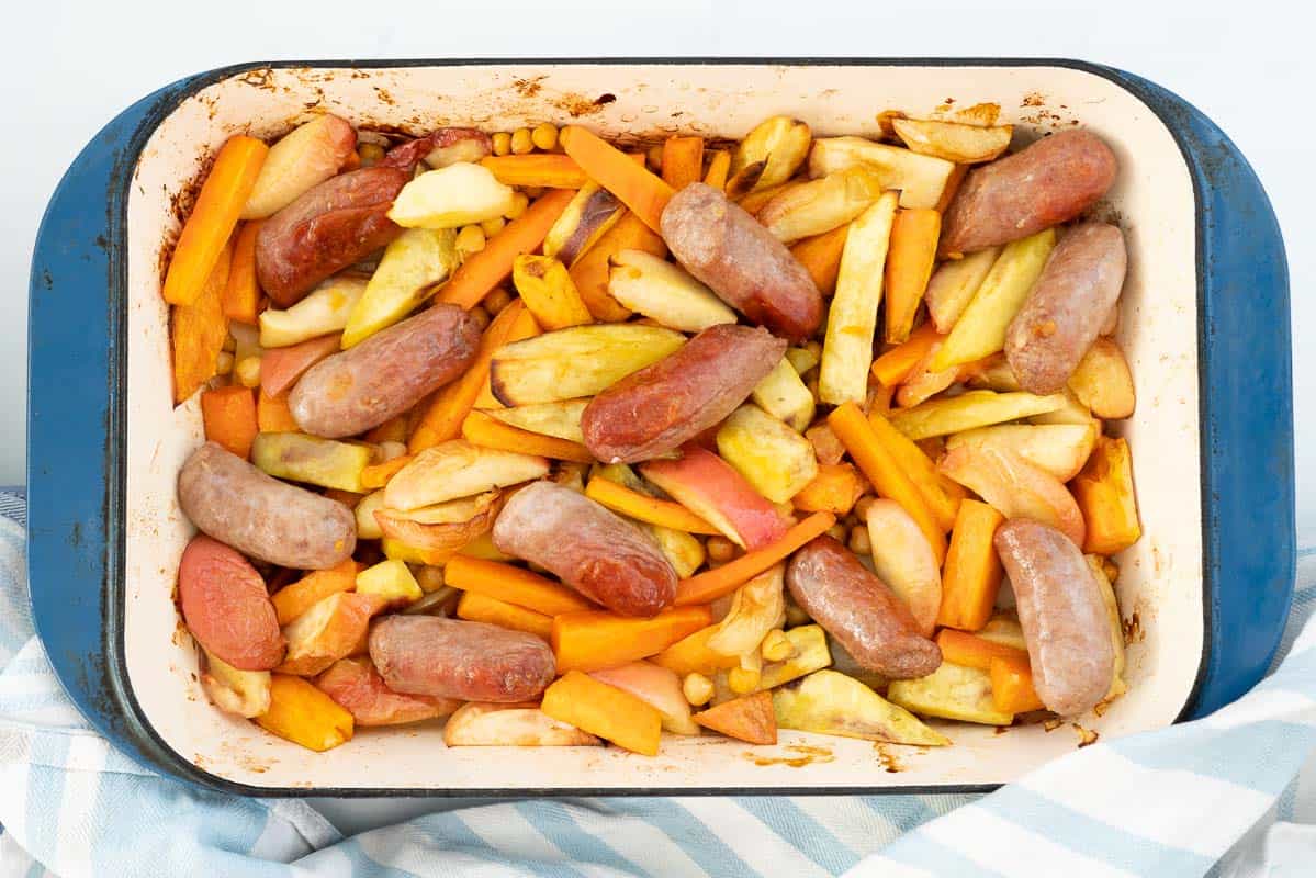 Cooked apples, carrots, sweet potato, sausages and chickpeas in a large blue ceramic roasting pan.