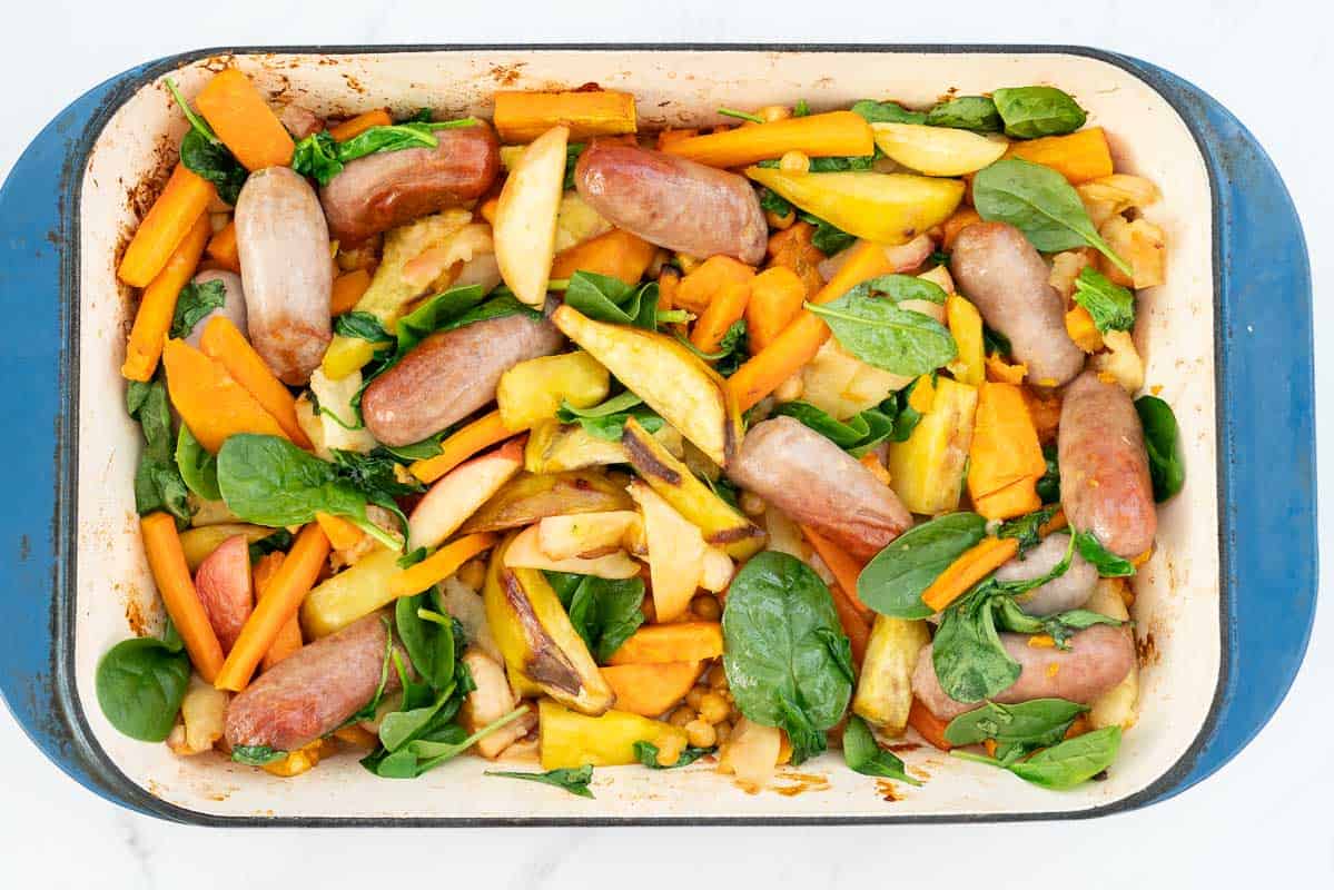 Cooked apples, spinach, carrots, sweet potato, sausages and chickpeas in a large blue ceramic roasting pan.