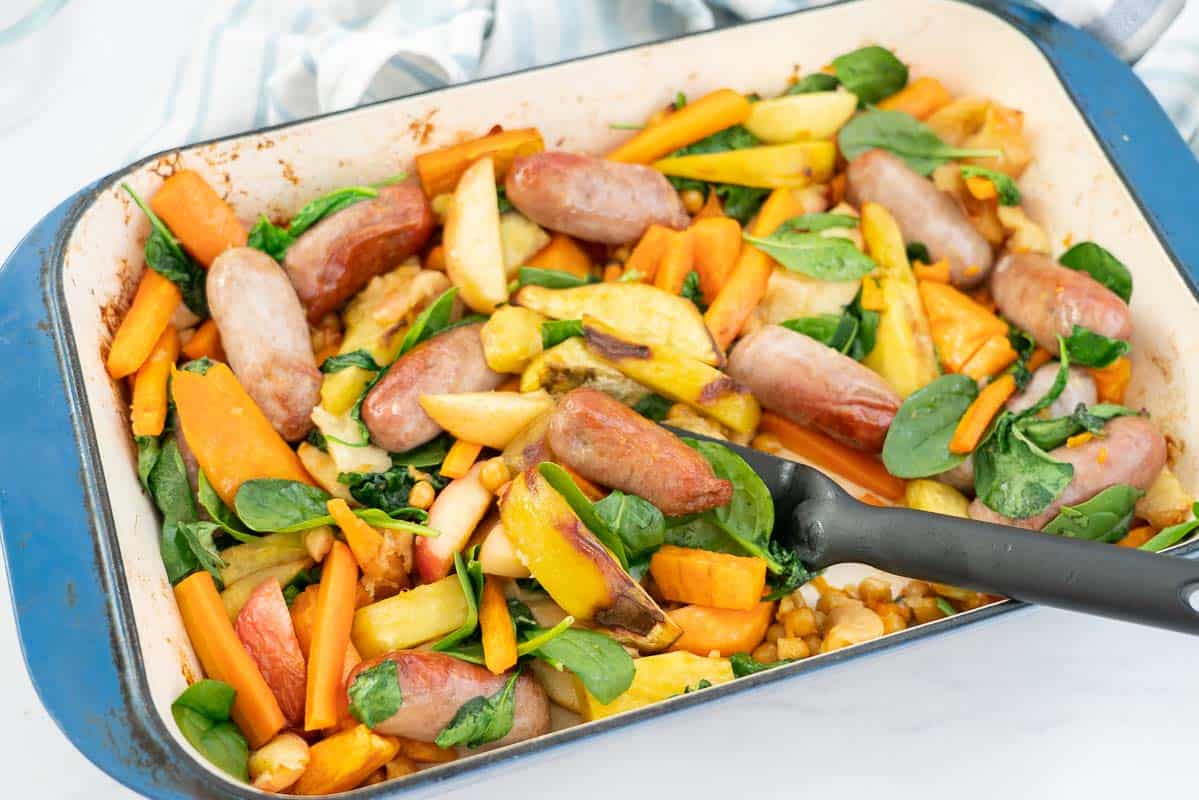 Baked sausages, apples, sweet potato and spinach leaves in a blue ceramic roasting pan.