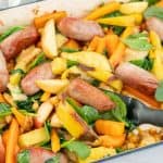 Baked sausages, apples, sweet potato and spinach leaves in a blue ceramic roasting pan.