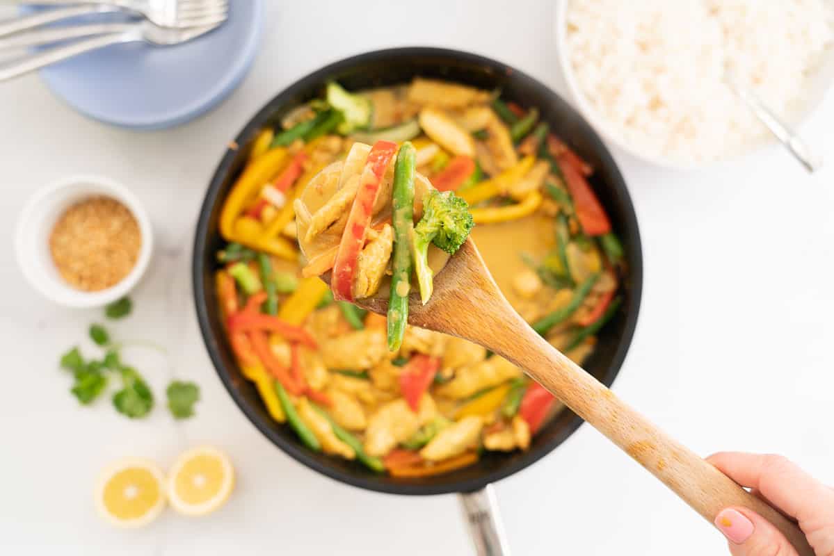 Wooden spoon of chicken satay stir fry being held above a large pan, rice and serving bowls.