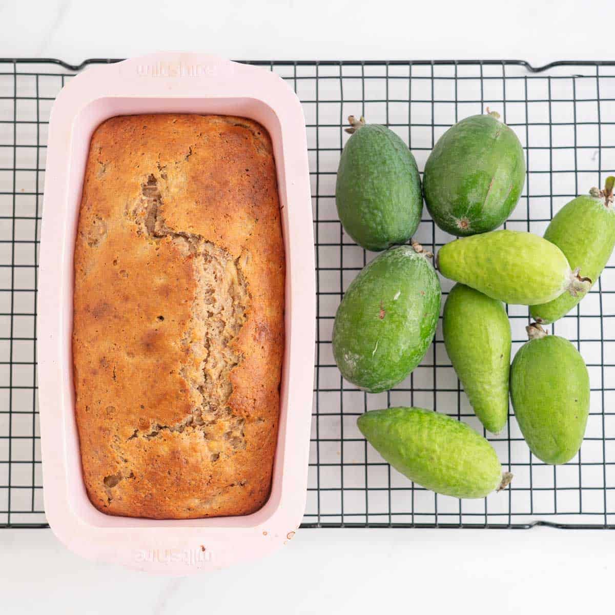 Golden brown loaf in a pink loaf tin, sitting on a cooling rack next to a pile of feijoas.