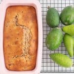 Golden brown loaf in a pink loaf tin, sitting on a cooling rack next to a pile of feijoas.