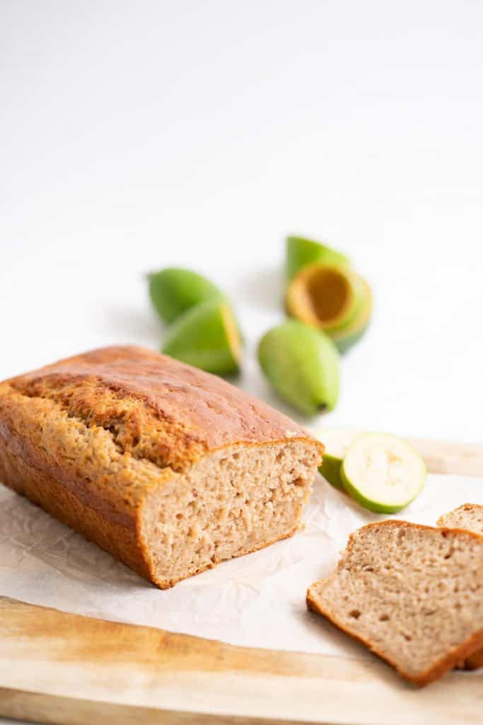 A feijoa loaf sitting on a wooden chopping board with 3 slices of loaf, bowl of fruit in the background.
