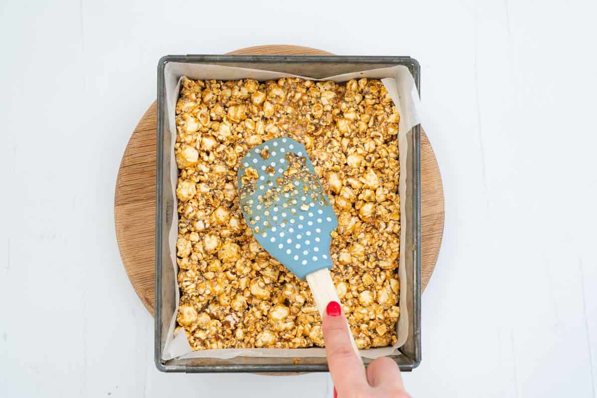 Caramel covered popcorn being pressed into a lined cake tin with a blue spatula.