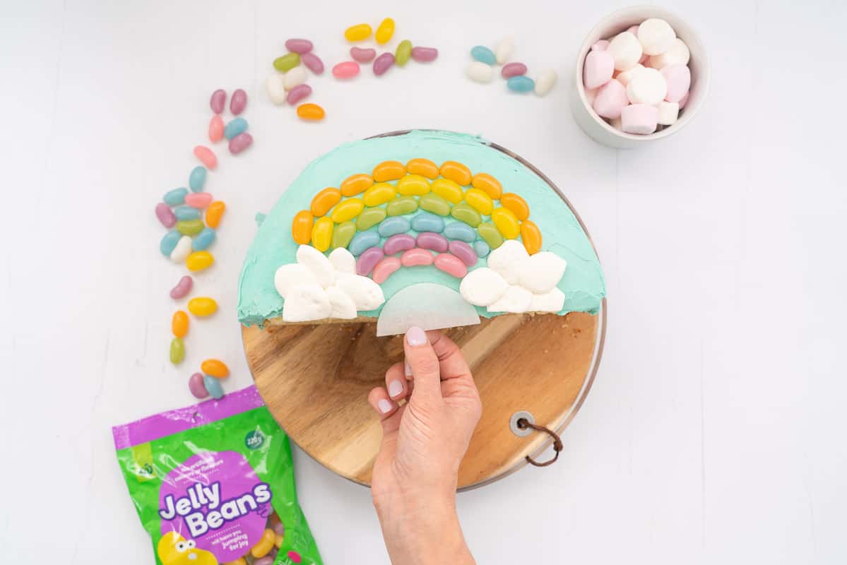 Half round blue cake decorated with a rainbow of jelly beans and marshmallow clouds, woman's hand removing the paper used as a guide for decoration placement.