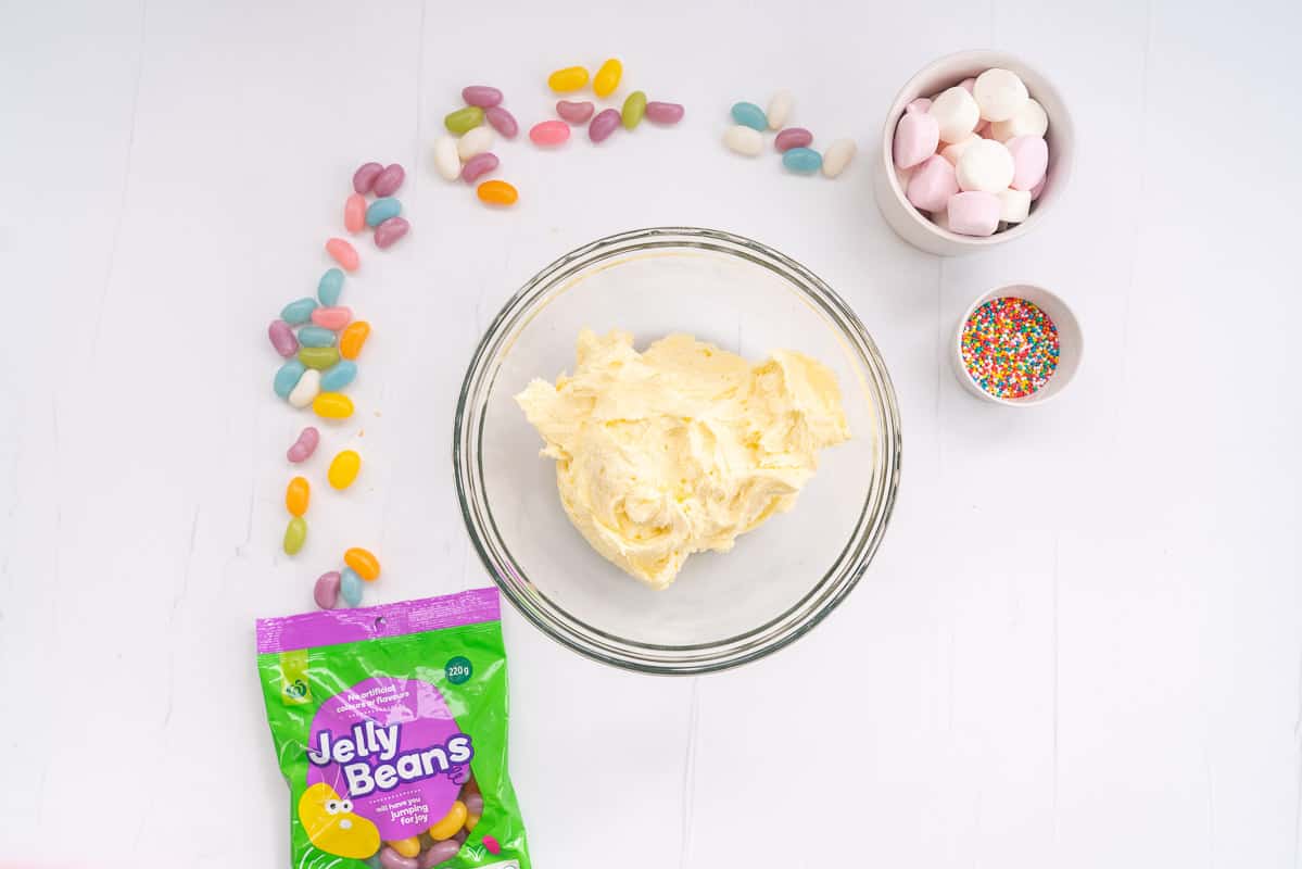 A glass bowl of buttercream on a bench with jelly beans, sprinkles and marshmallows.