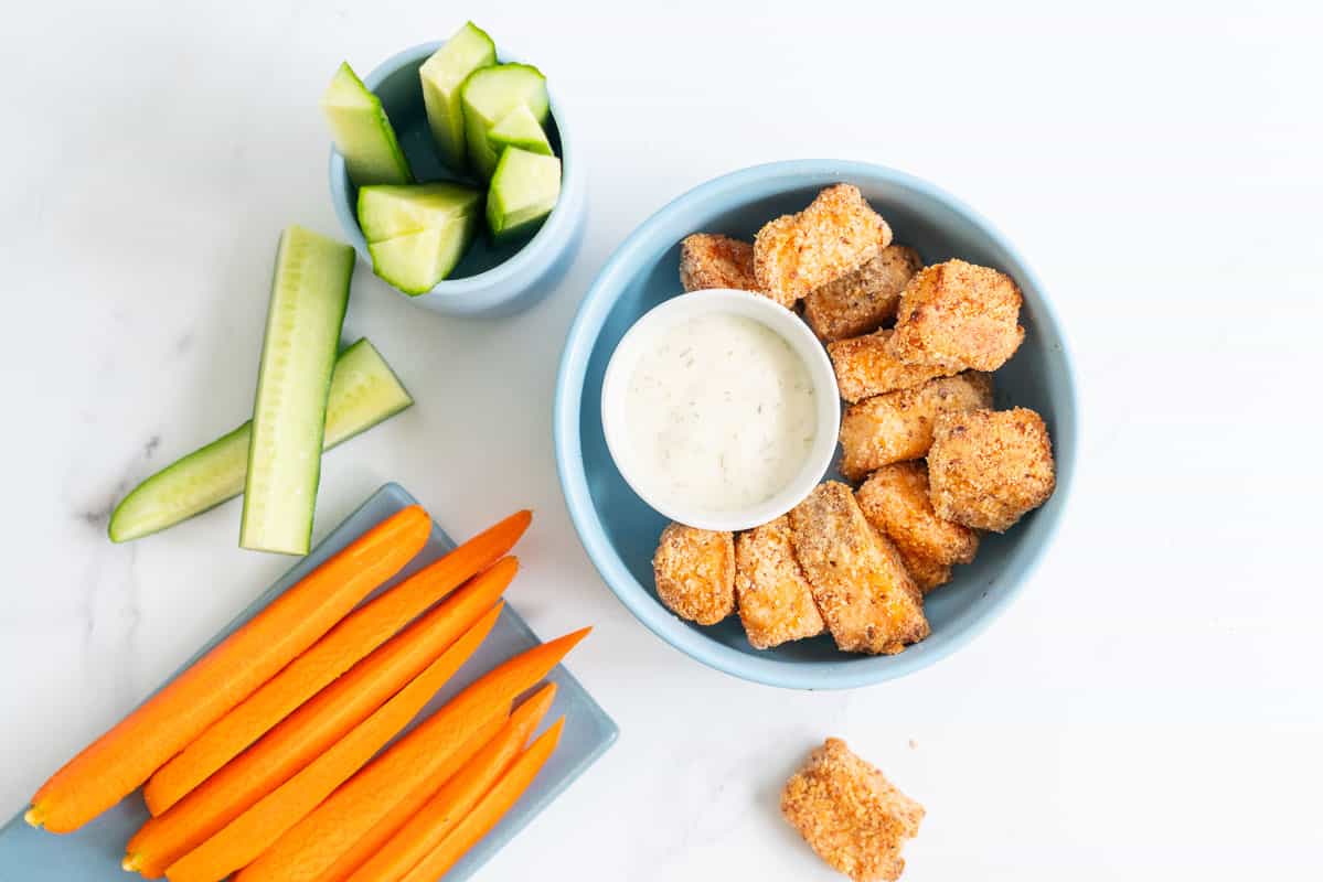 A blue bowl filled with salmon nuggets, alongside cucumber and carrot sticks.