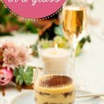 A tiramisu cup sitting on a table decorated with candles and fresh flowers, a glass of champagne, text overlay "Tiramisu in a Glass".