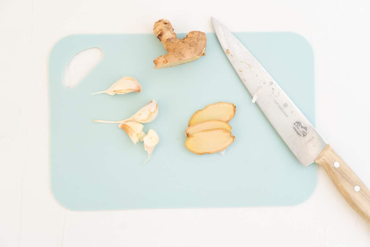 Slices of ginger and crushed garlic cloves on a light blue chooping board with a large chefs knife.
