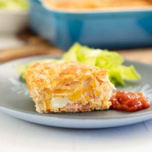 A slice of bacon and egg pie on a blue plate, with tomato relish and salad.