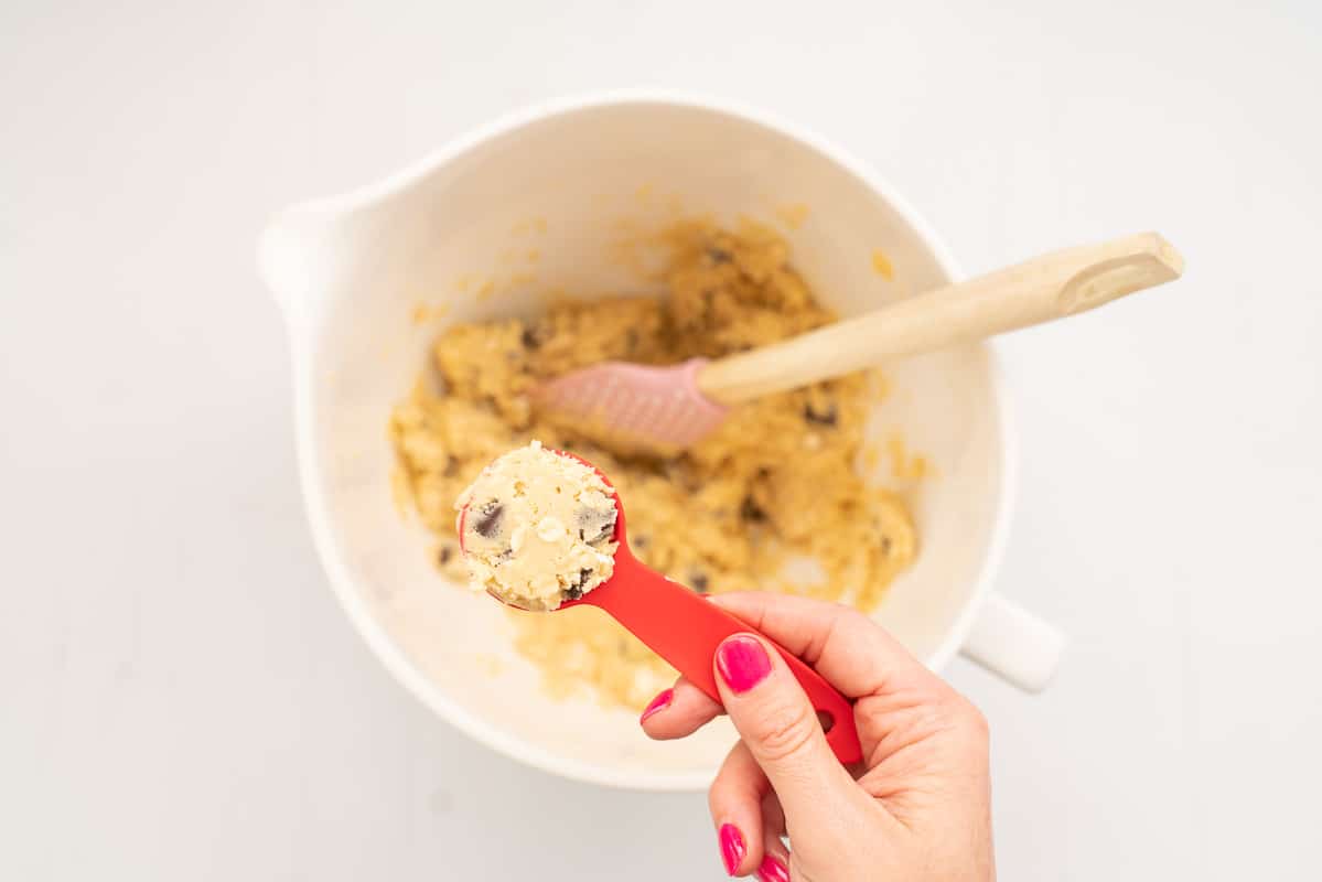 A women's hand holding a red tablespoon filled with cookie dough.