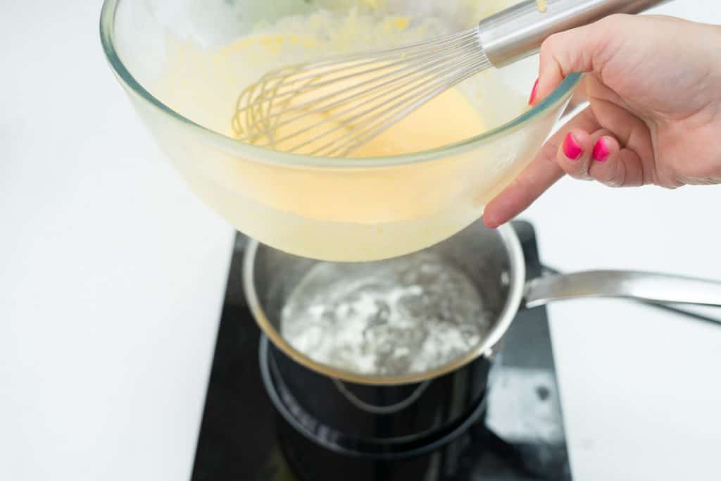 A hand holding a glass bowl of custard above a saucepan of boiling water. 
