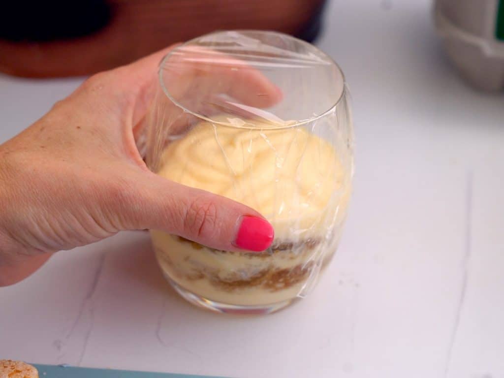 A women's hand picking up a Tiramisu cup covered in cling film ready to be refrigerated.