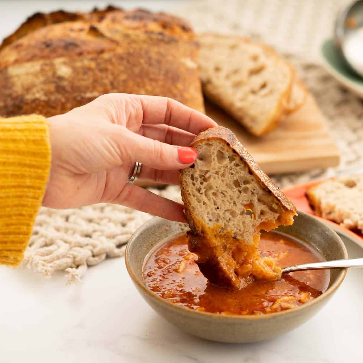 A woman's hand dipping a slice of crusty bread into a bowl of tomato soup.