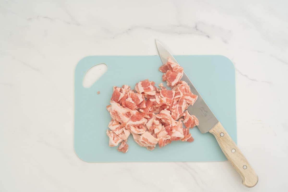 Diced streaky bacon on a light blue chopping board with a chefs knife.